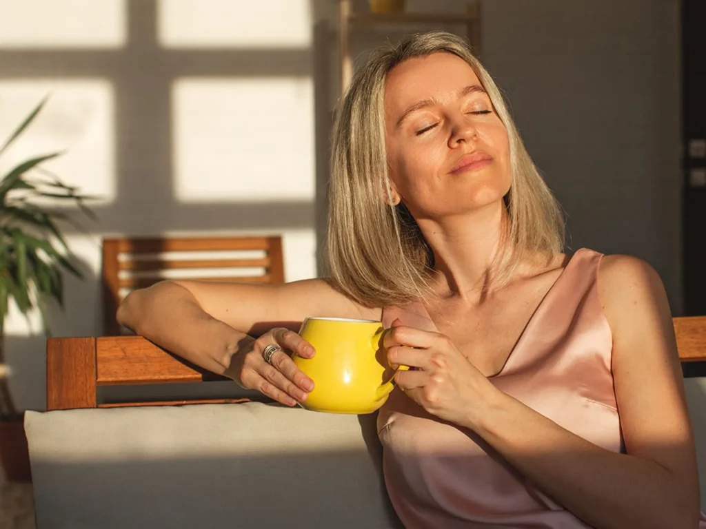 attractive happy middle aged woman is sitting on sofa in living room smiling adult lady enjoys drinking coffee or tea sitting o